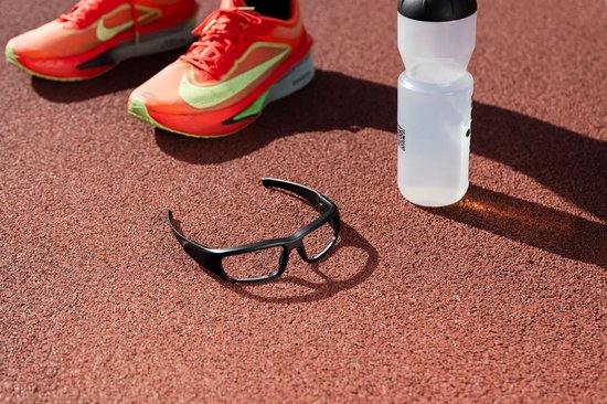 Zwarte bril op een rode atletiekbaan, naast sportschoenen en bidon.
