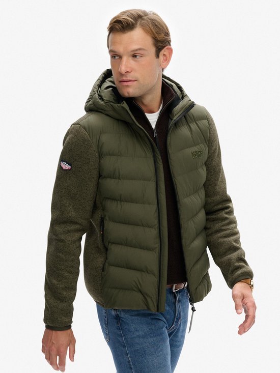 Man in groene jas met gewatteerd voorpand, capuchon en gebreide mouwen. Logo op de linker mouw. Draagt een bruine coltrui en spijkerbroek.