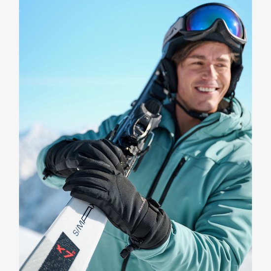 Man in een skibril en helm, draagt een blauwe jas en zwarte skihandschoenen, houdt ski's vast.