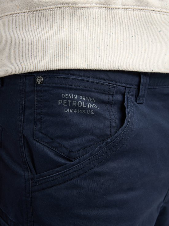 Sluiting van een donkerblauwe cargobroek met zakdetail en tekst: 'DENIM DRIVEN PETROL IND. DIV.4148-U.S.'.