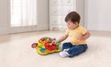 Een baby in een geel T-shirt en spijkerbroek zit op een lichte vloer naast een geel VTech stuurtje.