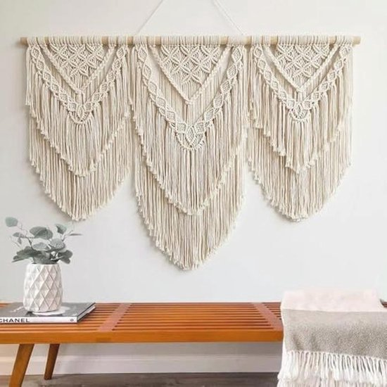 Crèmekleurig macramé wandkleed met drie V-vormige patronen, boven een houten bank met een vaas.