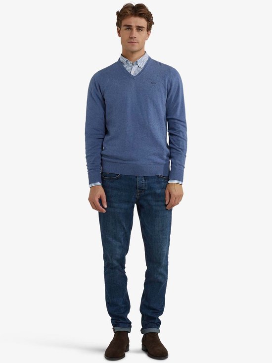 Man draagt een blauwe V-hals trui van merinowol over een gestreept overhemd, met jeans.