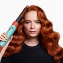 Vrouw met rood, golvend haar houdt Dyson Airwrap i.d. (blauw/oranje) vast.