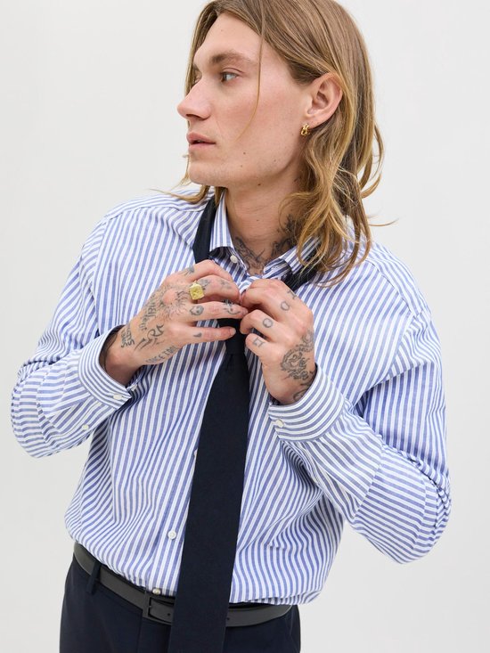 Man met lang blond haar, tatoeages, draagt een blauw-wit gestreept overhemd en knoopt een zwarte stropdas vast.