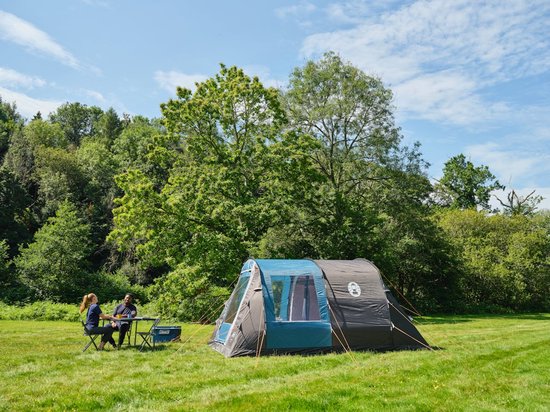 Grijze en blauwe tunneltent op gras met twee personen aan tafel. Bos op achtergrond.