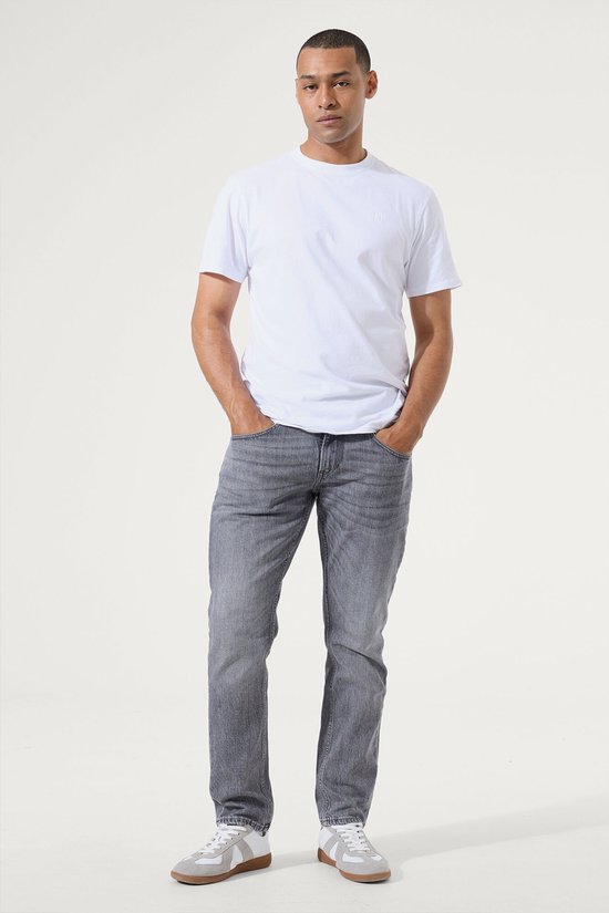 Man, met de handen in de zakken, draagt een grijze slim fit jeans met een witte t-shirt.