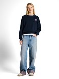 Model in een donkerblauwe sweater met een roze hartje en een wijde, lichtblauwe jeans met omgeslagen zoom.