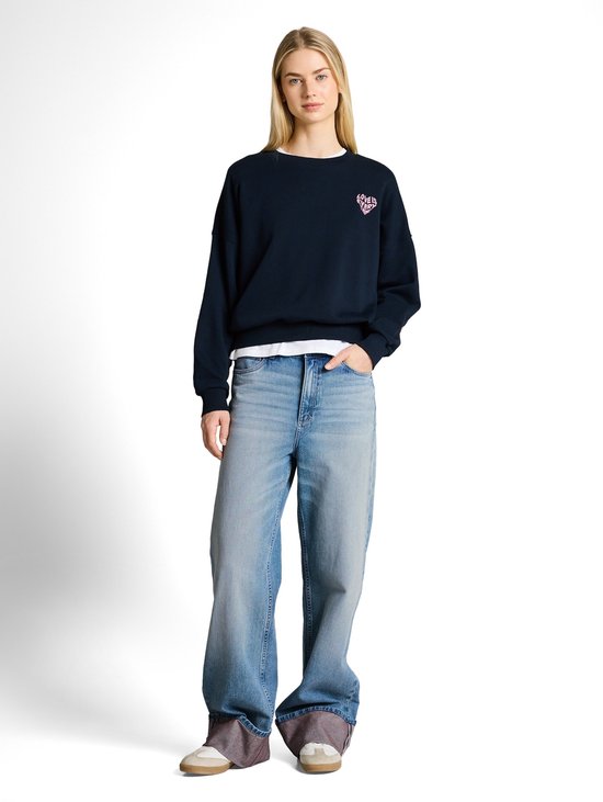Model in een donkerblauwe sweater met een roze hartje en een wijde, lichtblauwe jeans met omgeslagen zoom.