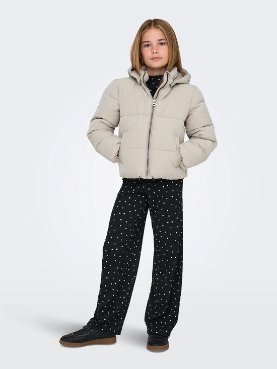 Meisje met blonde staartjes draagt een lichtbeige pufferjack met capuchon en zwarte broek met witte stippen.