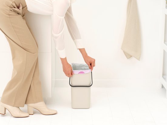 Vrouw doet afvalzak met strik in een licht beige prullenbak.