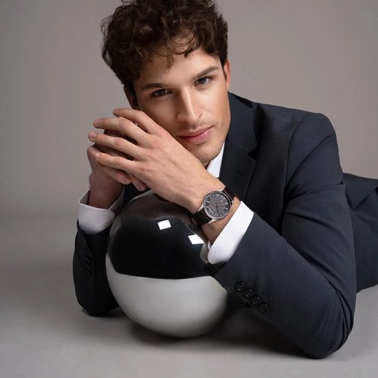 Man in donkergrijs pak met Jacques Lemans horloge om de pols, liggend op een bol.