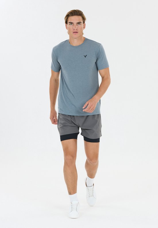 Man in blauw shirt en grijze sportshort met zwart binnenbroekje, witte sokken en witte schoenen.