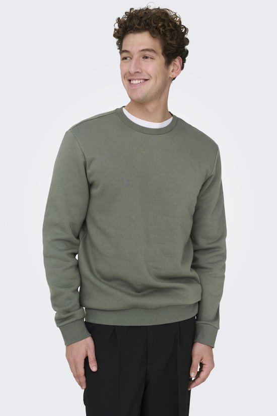 Een man met krullend haar draagt een mosgroene sweater met ronde hals en zwarte broek.
