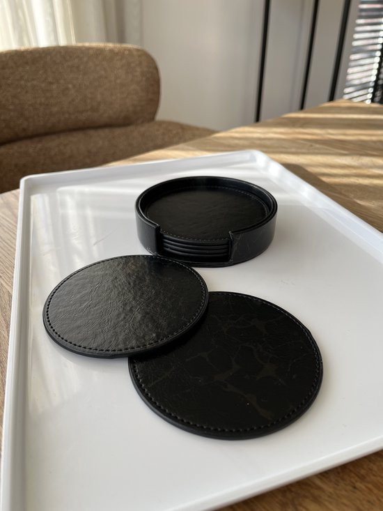 Zwarte ronde onderzetters met marmerprint en stiksels, op een witte schaal. Twee liggen los, de rest in een zwarte houder.