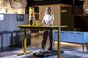 Vrouw staat op een AJ-Sports loopband voor een hoog-instelbaar bureau met laptop. De loopband is geplaatst op een vloerkleed.