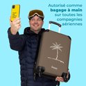 Man met skibril en muts neemt selfie met gele telefoon, houdt bruine koffer met palmboom op.