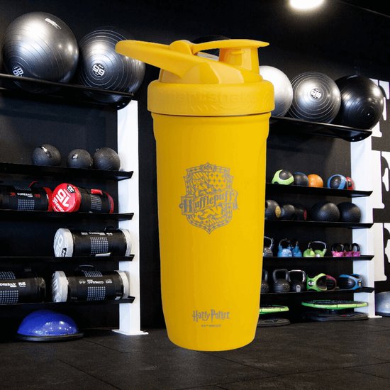 Gele Hufflepuff Smartshake-beker voor de fitness, met sportartikelen op de achtergrond.