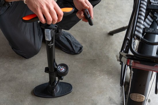 Persoon met horloge gebruikt zwarte fietspomp met oranje handvat en drukmeter naast een fietswiel.