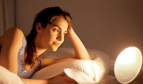 Een vrouw ligt op haar zij in bed, haar ogen zijn open en gericht op de wekker die zonsopkomst simuleert.