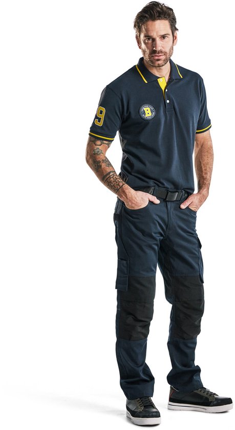 Man in donkerblauwe werkbroek en polo, staand met handen in zakken.