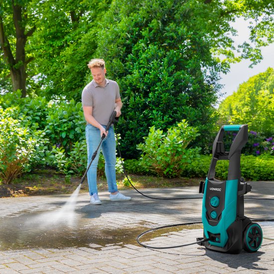Een man met grijze polo en spijkerbroek reinigt een terras met de groene en zwarte hogedrukreiniger.