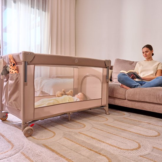 Een vrouw leest een boek naast een beige reisbedje met een slapende baby erin. Het reisbedje is bevestigd aan een ouderlijk bed.