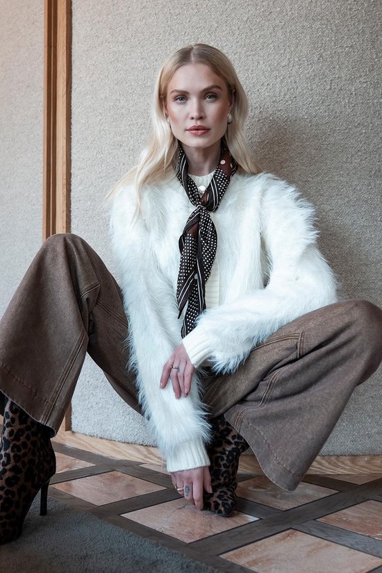 Model zittend in een off-white, harig vest, bruine broek, stippelsjaal en luipaardprint laarzen.
