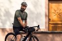 Man met olijfgroen fietsshirt, zwart fietsbroekje en helm, zittend op fiets.