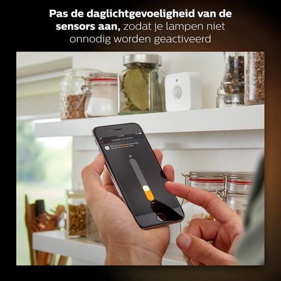 Een hand op een smartphone met de Hue app, die de daglichtgevoeligheid van de sensor aanpast. De witte sensor is zichtbaar op een schap.