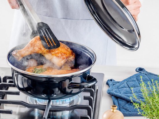 Een kipkluif wordt met een tang uit een zwarte braadpan gehaald, damp komt uit de pan. De pan staat op een gasfornuis.
