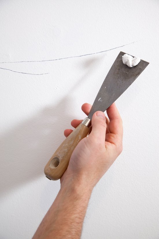 Hand met spatel en witte houtrotvuller, brengt vuller aan op witte muur.