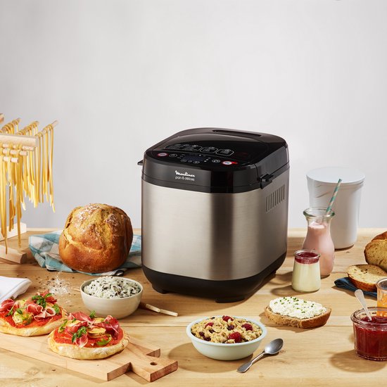 Zilverkleurige broodbakmachine op een houten tafel, omringd door diverse broden, pasta, beleg en ontbijtproducten.