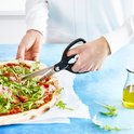 Zwarte keukenschaar snijdt een pizza met ham en rucola op een vel bakpapier.