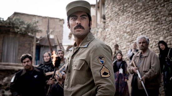 Een man met snor in beige militair uniform en pet, kijkt weg met een muur van stenen achter zich.