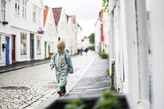 Jongen in groen gestreept regenpak loopt op geplaveide straat in een stadje.