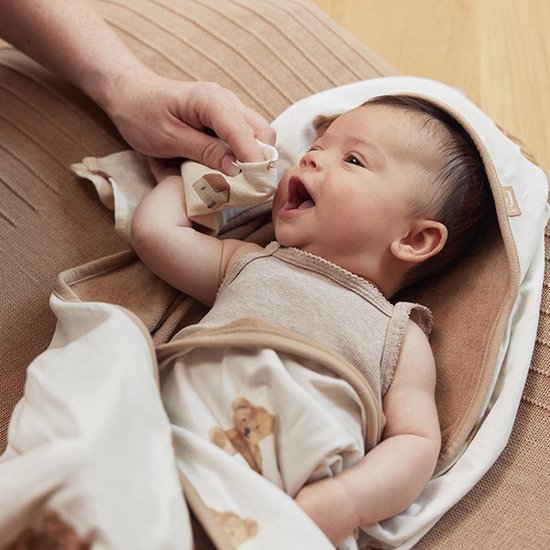 Un bébé souriant allongé dans une cape de bain. Une main tend un gant de toilette avec un imprimé d'ours brun au bébé.