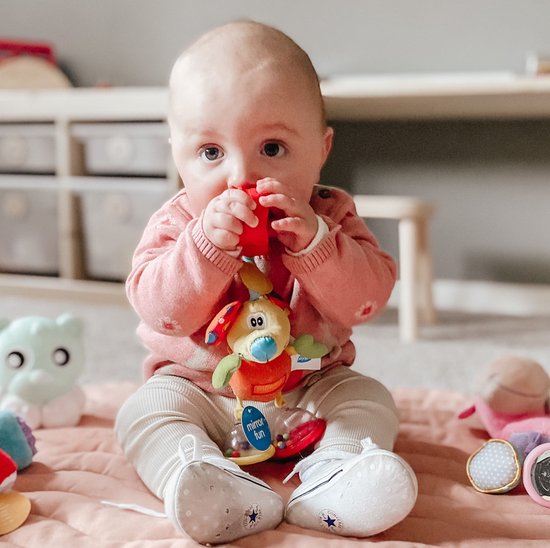 Een baby zit op een kleed en pakt Mimsy de Muis, een gele knuffel met oranje lijf, groene armen en rode oren met stippen.