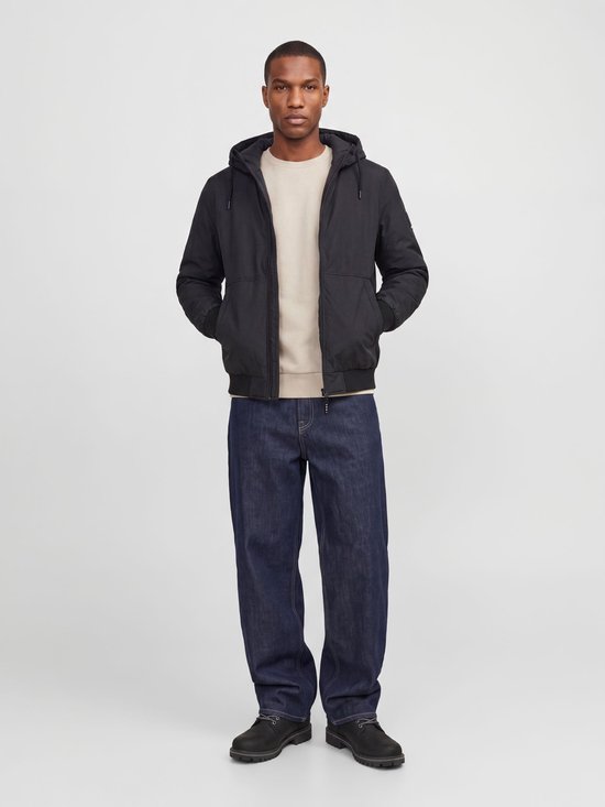 Man met crème kleurige sweater, zwarte jas met capuchon en donkerblauwe jeans. De jas is open.
