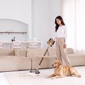 Vrouw met bruin haar in witte top en beige broek zuigt met gouden steelstofzuiger, golden retriever ligt naast haar.
