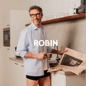 Man met bril en krant in de keuken, draagt een lichtblauwe blouse en donkerblauwe Robin boxershorts.