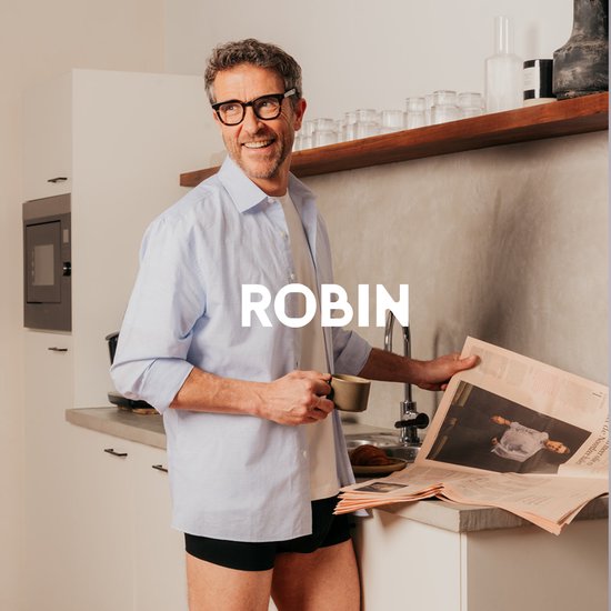 Man met bril en krant in de keuken, draagt een lichtblauwe blouse en donkerblauwe Robin boxershorts.