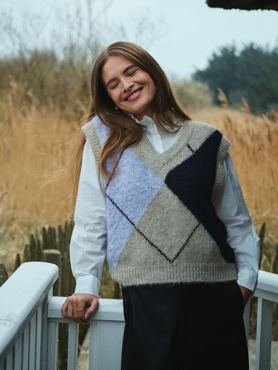 Model in een beige gebreide trui met argyle patroon in paars en marineblauw, gedragen over een wit overhemd.