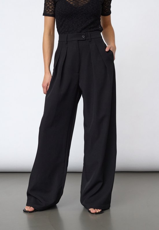 Model met zwarte pantalon met brede pijpen, plooien bij de taille en zwarte kanten top.