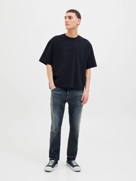 Man in zwart oversized T-shirt en donkerblauwe jeans met grijze wassing, handen in zakken.