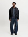 Man draagt een donkerblauwe poloshirt, zwarte jas en jeans.