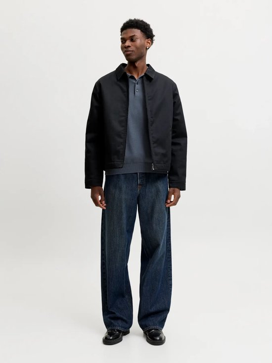 Man draagt een donkerblauwe poloshirt, zwarte jas en jeans.