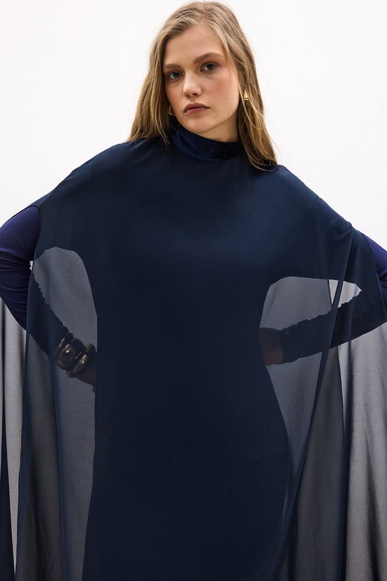Donkerblauwe jurk met hoge hals en wijde, doorschijnende mouwen van chiffon over een nauwsluitende, lange mouw.