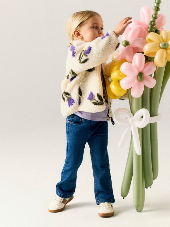 Meisje in een spijkerbroek met rechte pijpen en een beige teddy jas met paarse bloemenprint, bij een ballonboeket.