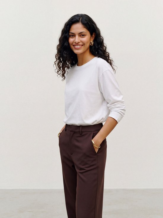 Lachend model draagt een wit shirt met lange mouwen en een donkerbruine pantalon, handen in de zakken.